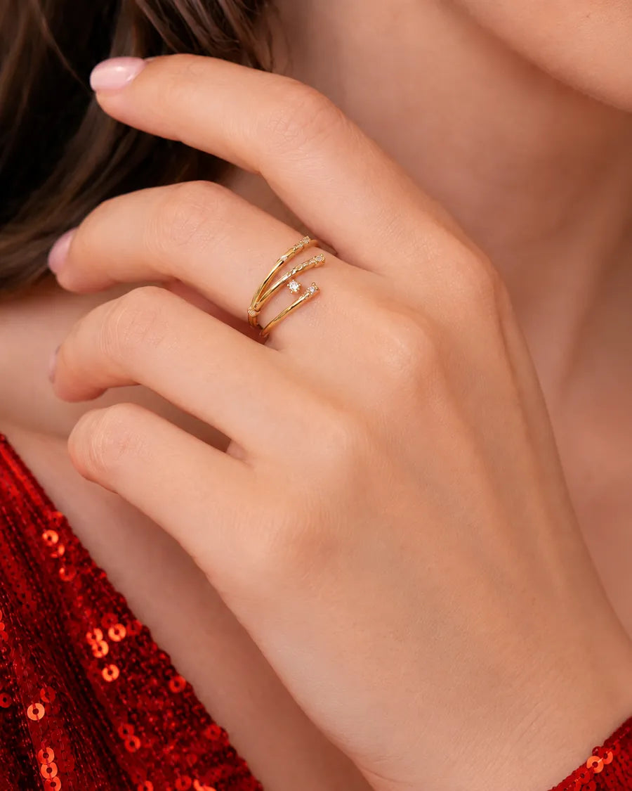 A close-up of a person's hand wearing two delicate gold wrap rings, with a small clear stone accent on one band. The person is wearing a red sequined top, and the background is softly blurred.